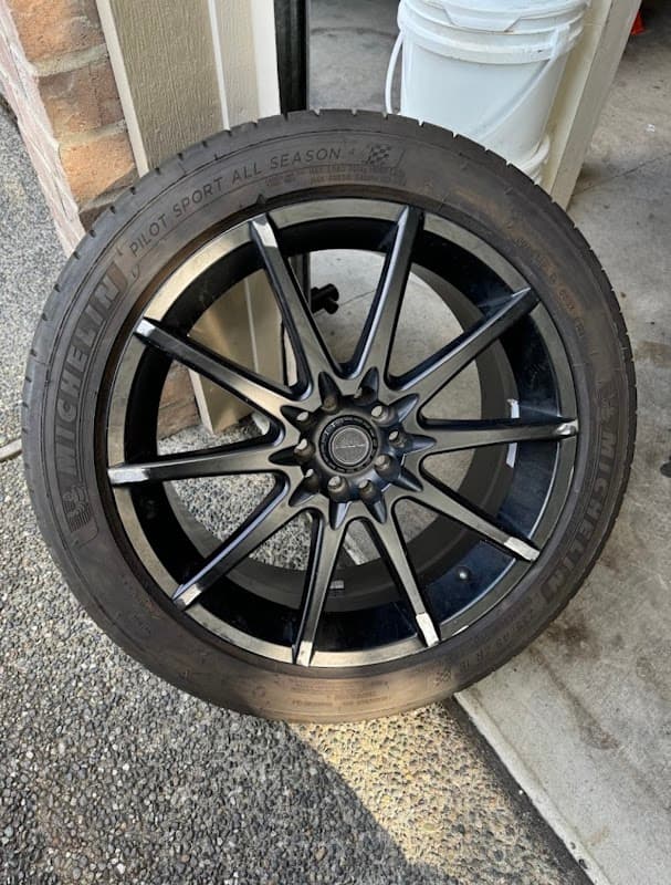 A Michelin Pilot Sport All Season tire mounted on a black multi-spoke alloy rim, stood outside the shop.